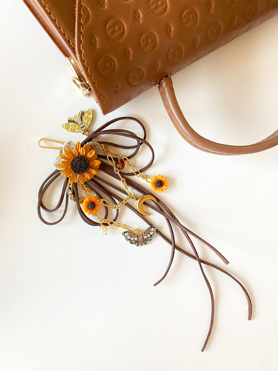 A brown leather handbag accessory with long ribbon-like strands, a sunflower charm, small orange flower charms, gold-toned details, and butterfly motifs, displayed on a white surface next to a brown embossed handbag.