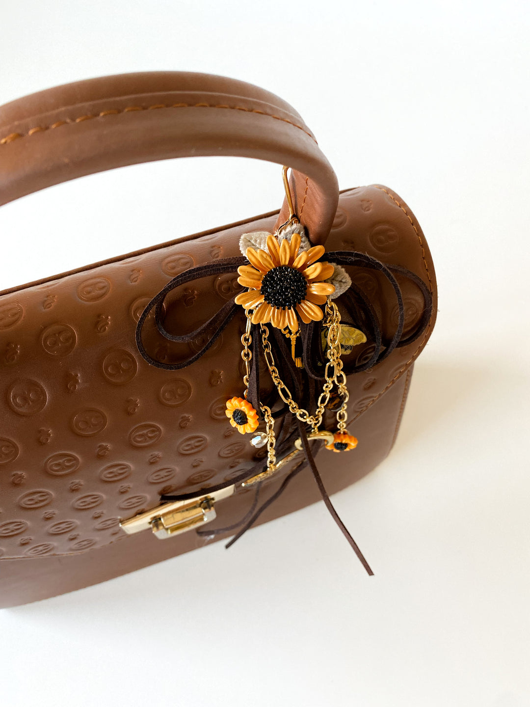 A brown textured leather handbag with a top handle, decorated with a dangling sunflower charm featuring gold chains and small orange accents, photographed on a clean white background.