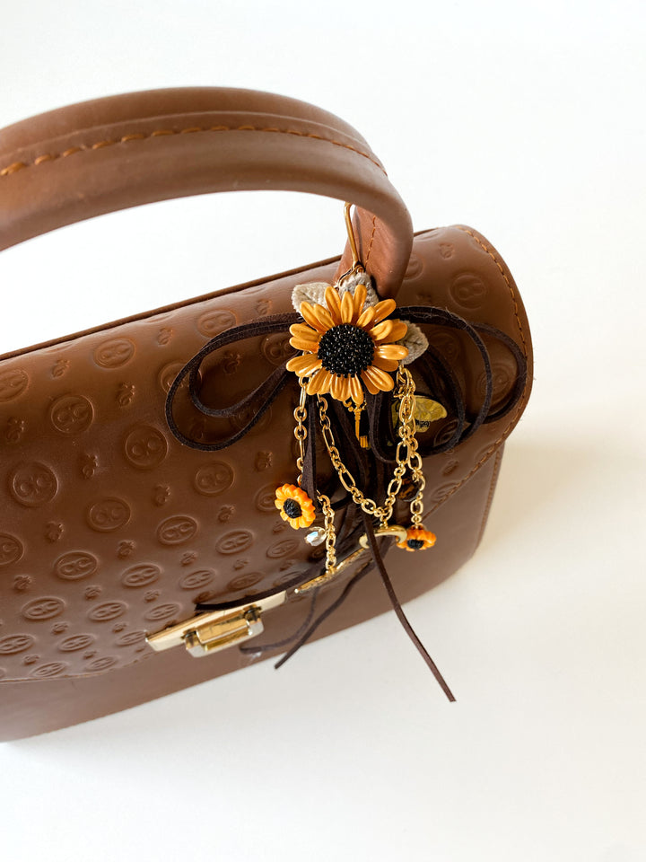 A brown textured leather handbag with a top handle, decorated with a dangling sunflower charm featuring gold chains and small orange accents, photographed on a clean white background.