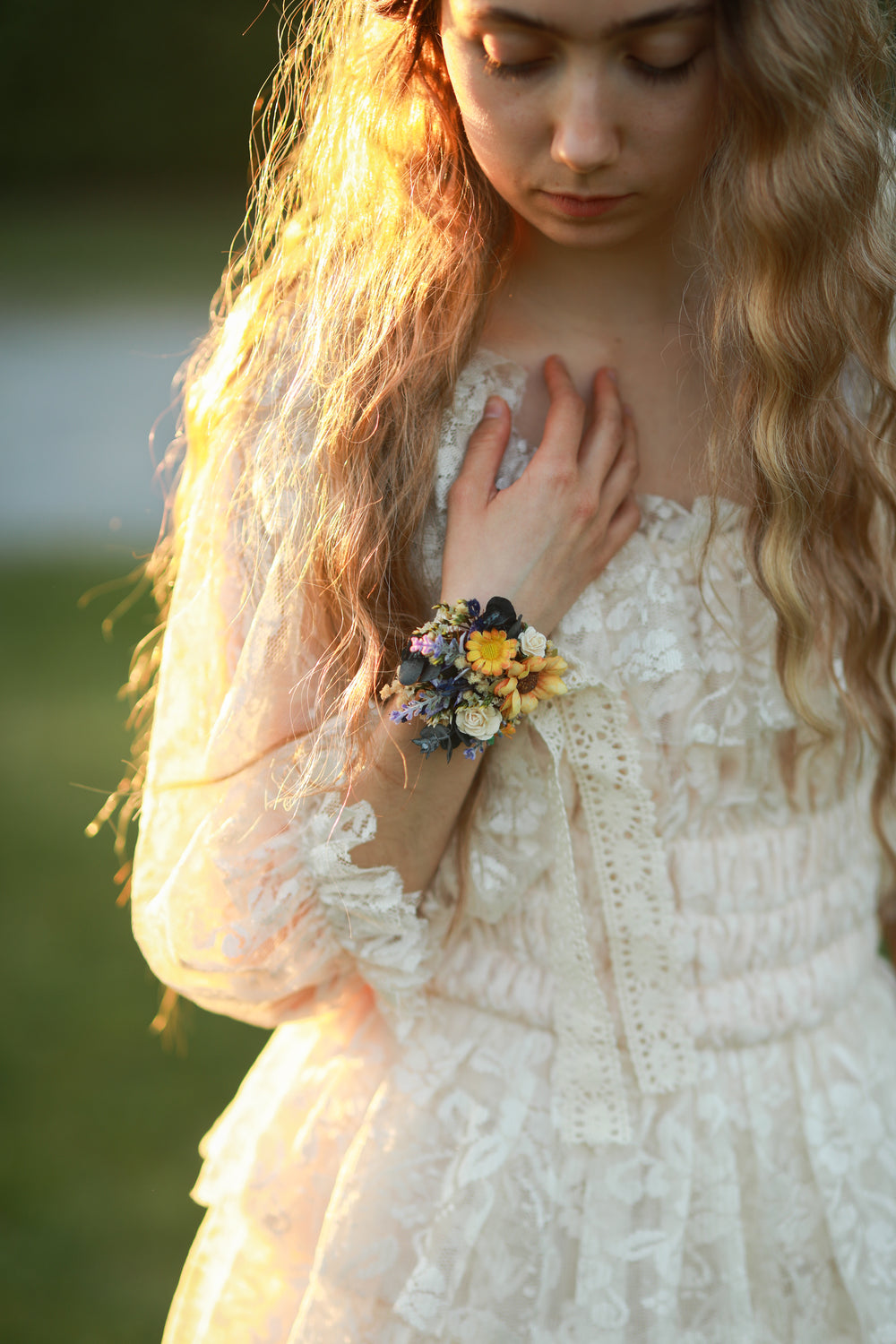 Woman in a white lace dress with a floral corsage, standing outdoors with sunlight.