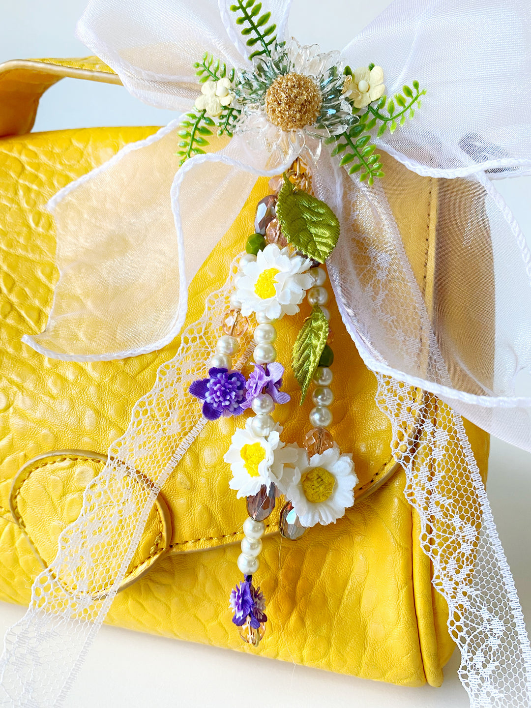 A close-up of a bright yellow textured handbag decorated with a large white tulle bow, white lace trim, and a dangling floral charm featuring white daisies with yellow centers, purple flowers, green leaves, and pearl beads.
