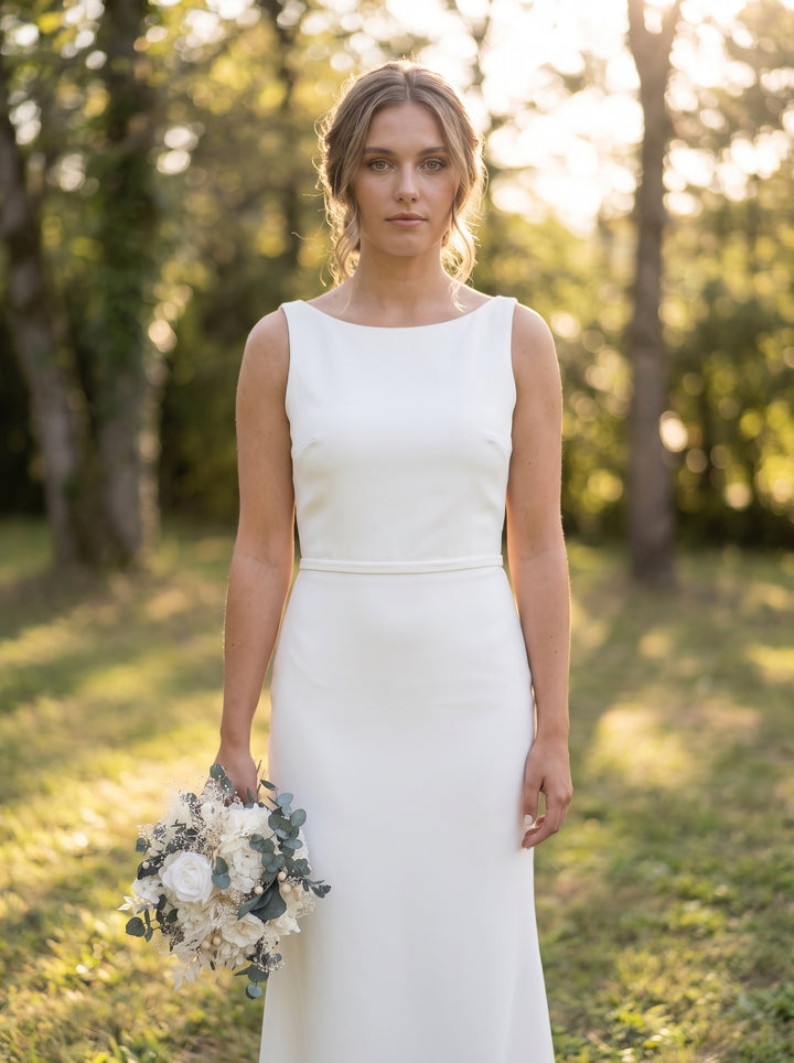 A woman wearing a sleeveless white wedding dress stands outdoors in a sunlit wooded setting, holding a bouquet of white and pale flowers.