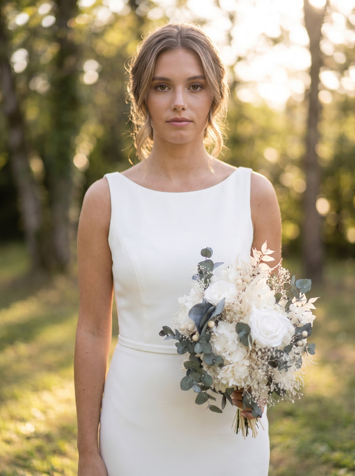 A woman wearing a sleeveless white wedding dress stands outdoors in a sunlit wooded setting, holding a bouquet of white and cream flowers with green foliage.