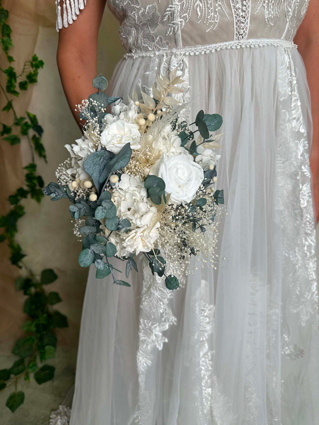 A close-up of a person wearing a white lace and tulle wedding dress while holding a bouquet of white flowers, green eucalyptus leaves, and small pearl-like accents.