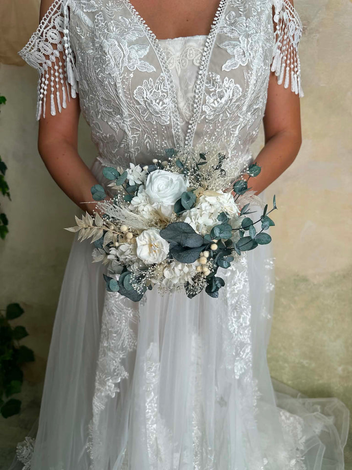 A bride wearing a white lace wedding dress with tassel fringe on the sleeves, holding a white and green  floral bouquet against a neutral textured wall.