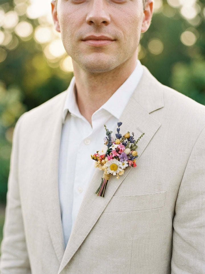 A close-up of a man wearing a light beige linen suit jacket over a white collared shirt, featuring a small colorful wildflower boutonniere on his lapel against a soft green bokeh background.