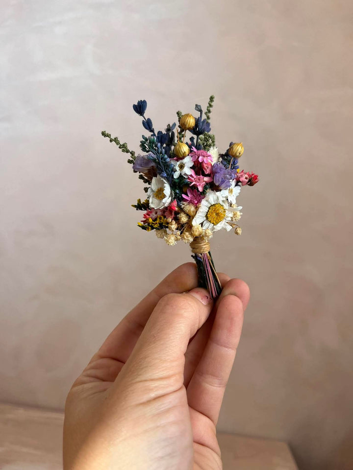 A hand holding a small dried flower boutonniere with white daisy-like flowers, purple blossoms, blue sprigs, and gold-toned buds against a soft beige wall background.