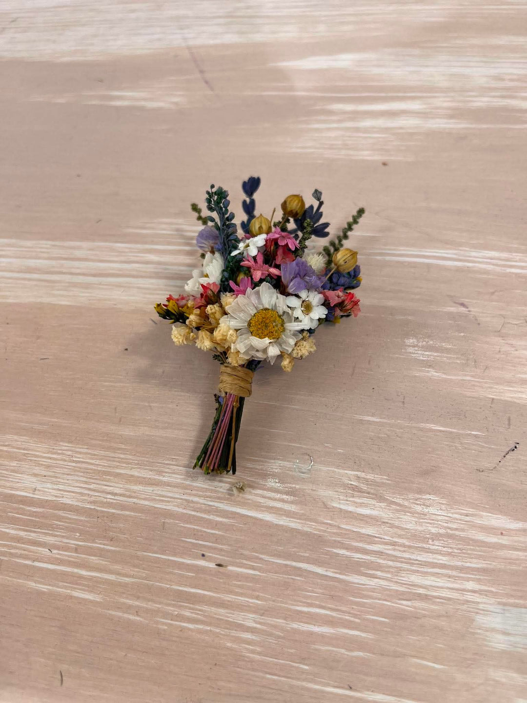A small dried flower boutonniere with white daisy-like blooms, yellow centers, pink and purple accents, and green stems, wrapped in tan paper and tied with multicolored string, placed on a light wood surface.
