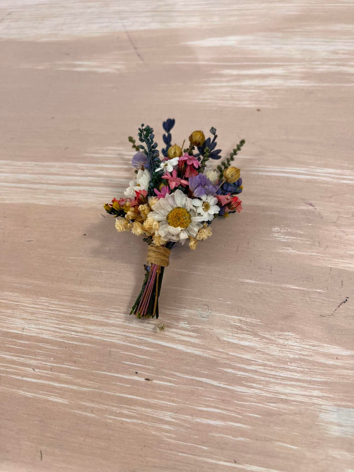 A small dried flower boutonniere with white daisy-like blooms, yellow centers, pink and purple accents, and green stems, wrapped in tan paper and tied with multicolored string, placed on a light wood surface.