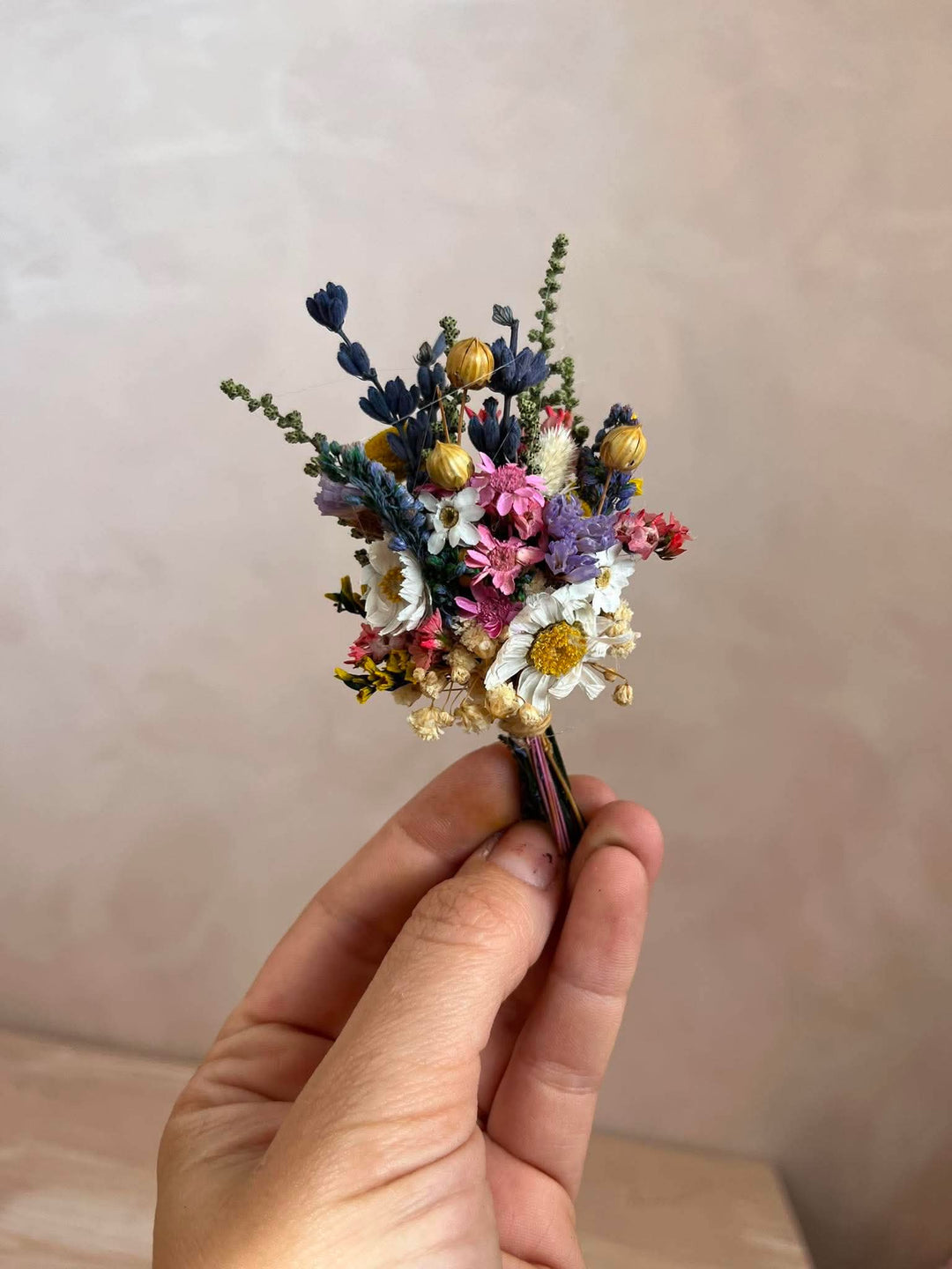 A hand holding a small dried flower boutonniere with white daisies, pink and purple blossoms, blue sprigs, and small gold-toned buds against a soft beige wall background.