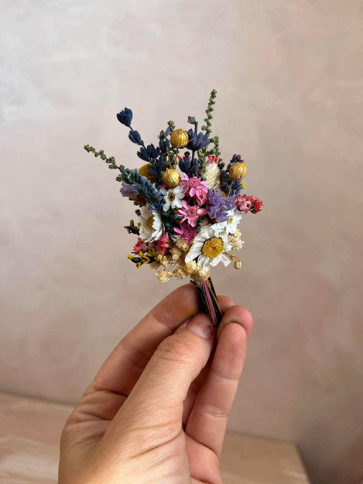 A hand holding a small dried flower boutonniere with white daisies, pink and purple blossoms, blue sprigs, and small gold-toned buds against a soft beige wall background.