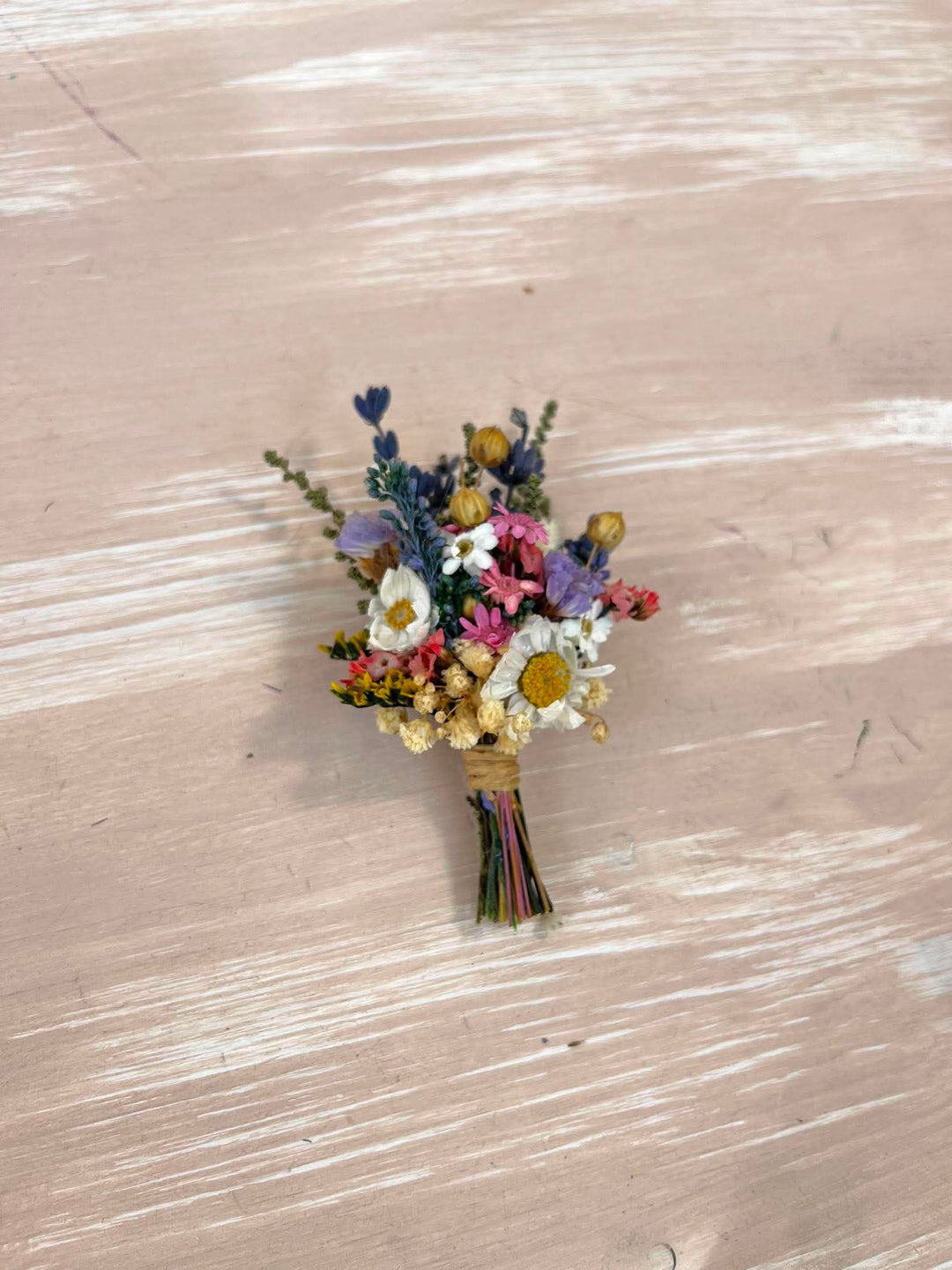 A small dried flower boutonniere with white daisies, pink blossoms, purple flowers, and yellow centers, wrapped in twine and placed on a light beige wooden surface.
