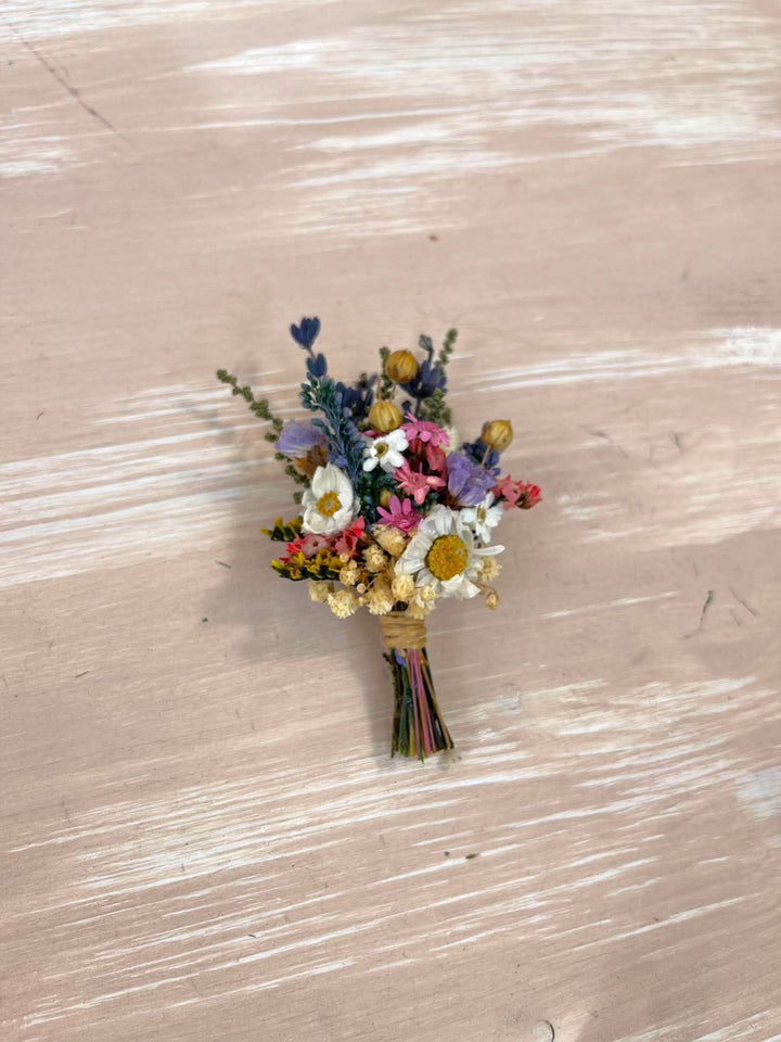 A small dried flower boutonniere with white daisies, pink blossoms, purple flowers, and yellow centers, wrapped in twine and placed on a light beige wooden surface.