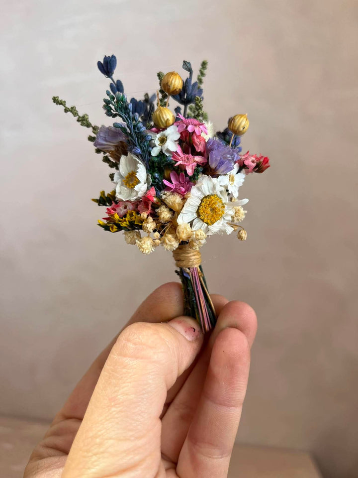 A hand holding a small dried flower boutonniere with white daisies, purple and pink blossoms, blue sprigs, and gold-toned buds wrapped in twine.