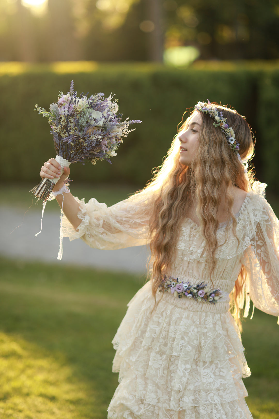 Woman in a vintage-style dress holding a bouquet, crown and belt of flowers outdoors.