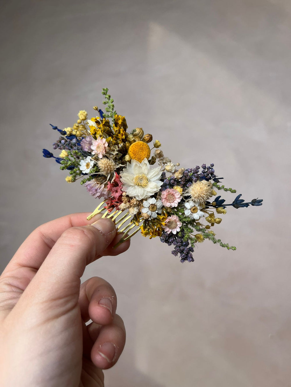 Hand holding a floral hair comb from dried flowers against a neutral background