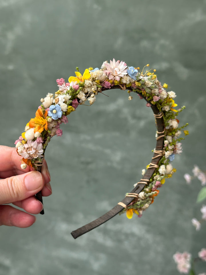A handmade floral headband wrapped with small flowers and greenery, held by a hand against a textured gray-green background.