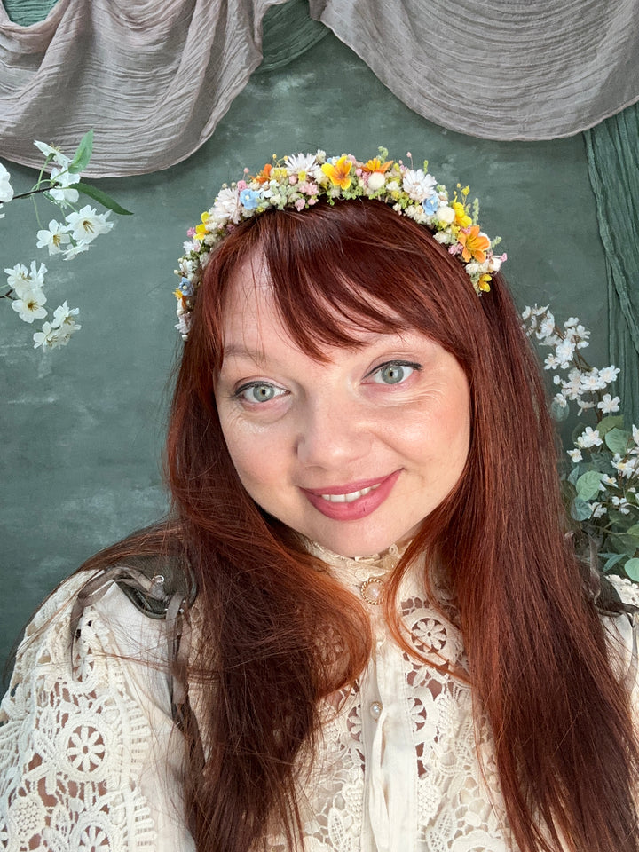 A smiling woman with long red hair wearing a colorful wildflower crown, posed against a dark green backdrop with draped fabric and white flowers.