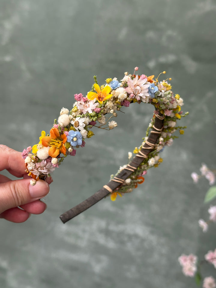 A handmade floral headband decorated with small flowers in pink, yellow, blue, and white, wrapped with twine on a dark wooden band, held by a hand against a textured gray background.