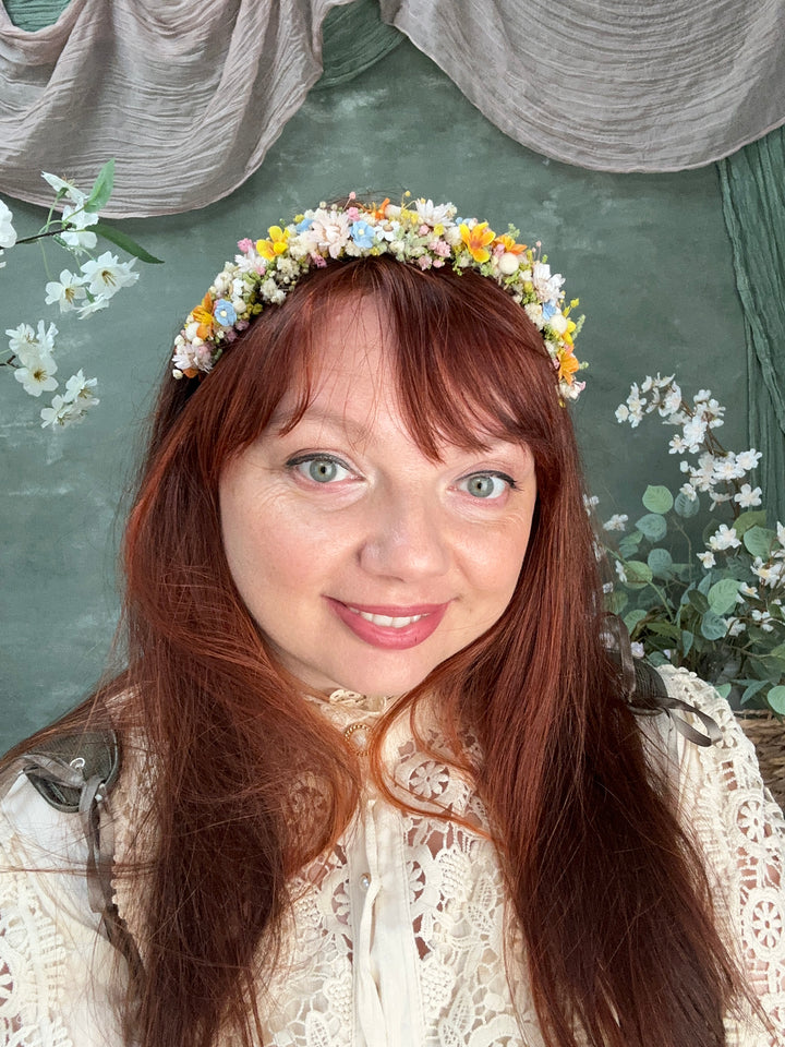 A woman with long red hair wearing a delicate white lace top and a colorful flower crown, lying on a textured green surface with eucalyptus leaves.