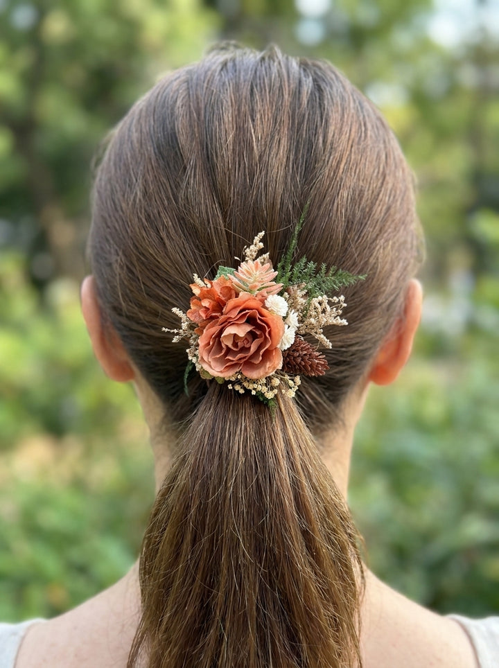 A woman viewed from behind with her hair in a low ponytail, wearing a decorative floral hair accessory with flowers and greenery at the base of the ponytail. The background is a softly blurred outdoor green setting.