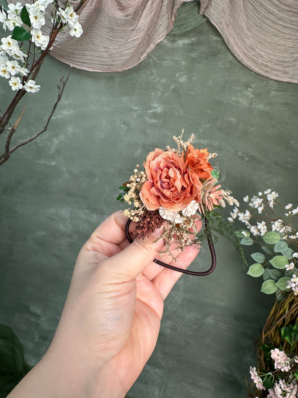 A hand holds a smal hair tie accessory with peach and orange flowers, beige filler stems, and green foliage against a textured sage green backdrop with decorative branches and draped fabric.