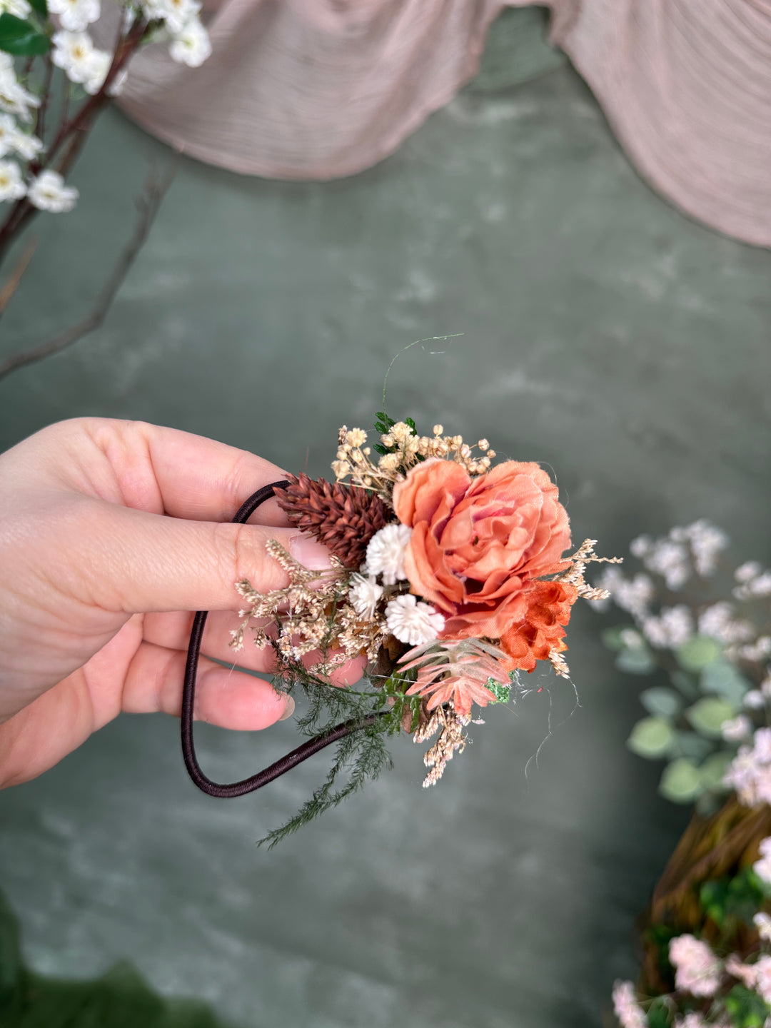A hand holding a s hair tie with a peach-orange rose, baby's breath, pinecone, and greenery attached to a dark elastic band, against a soft gray-green textured background.