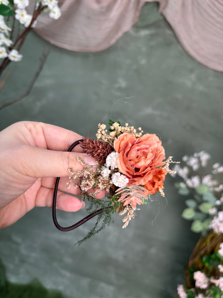 A hand holding a s hair tie with a peach-orange rose, baby's breath, pinecone, and greenery attached to a dark elastic band, against a soft gray-green textured background.