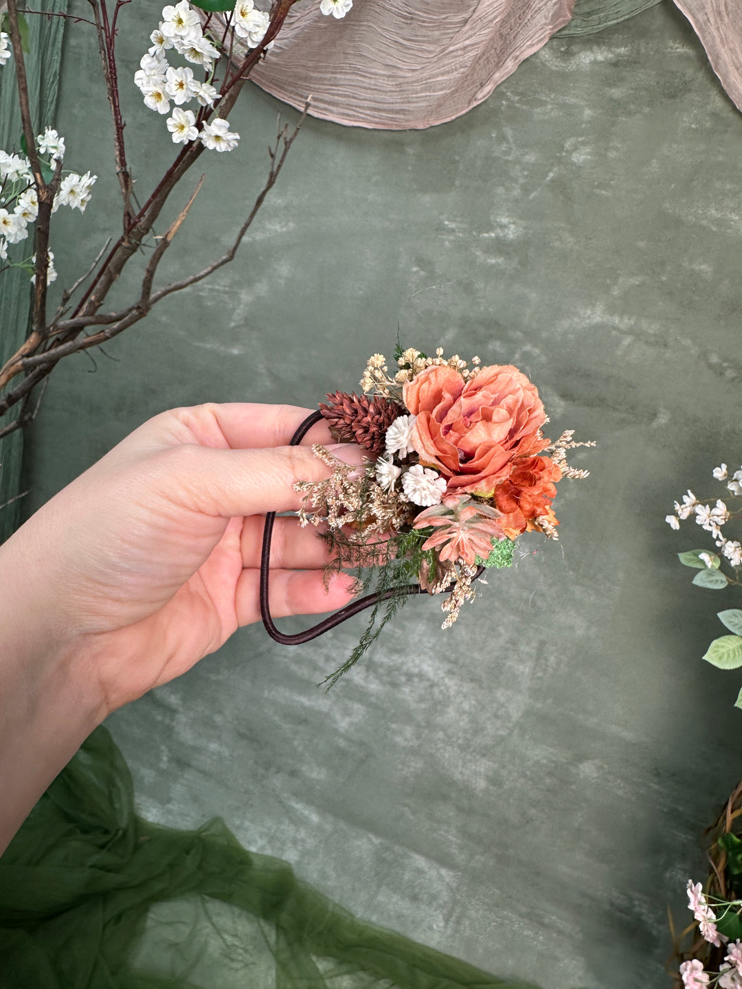 A hand holds a small floral hair accessory with peach and coral roses, white flowers, green leaves, and brown pinecones against a textured sage green backdrop with decorative branches.