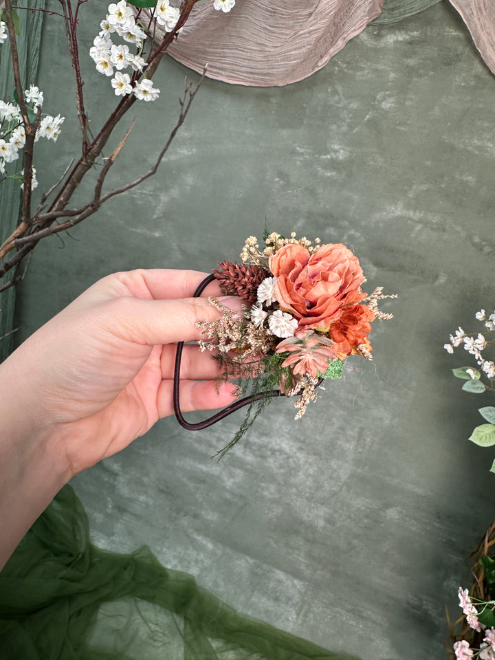 A hand holds a small floral hair accessory with peach and coral roses, white flowers, green leaves, and brown pinecones against a textured sage green backdrop with decorative branches.