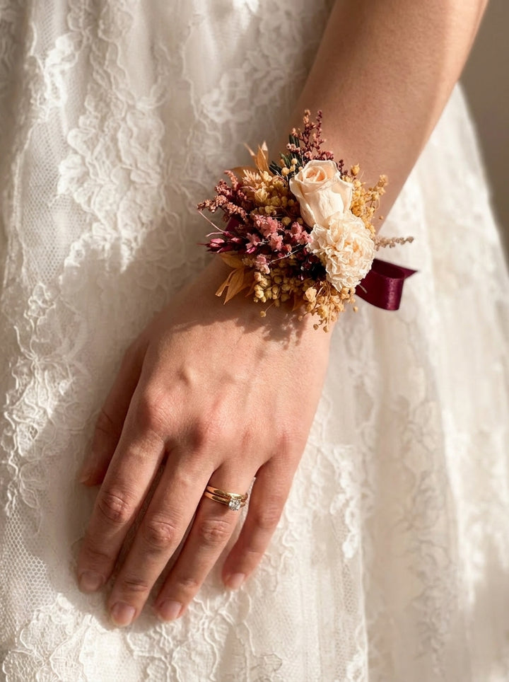 A close-up of a person's hand and wrist wearing a dried floral wrist corsage with cream, pink, and burgundy tones, set against a white lace wedding dress.