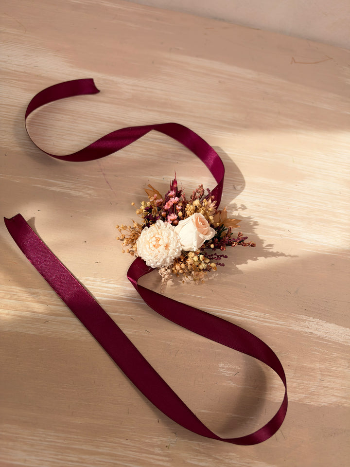 A dried floral bracelet with cream and burgundy flowers, tied with a long, flowing burgundy satin ribbon on a light wood surface.