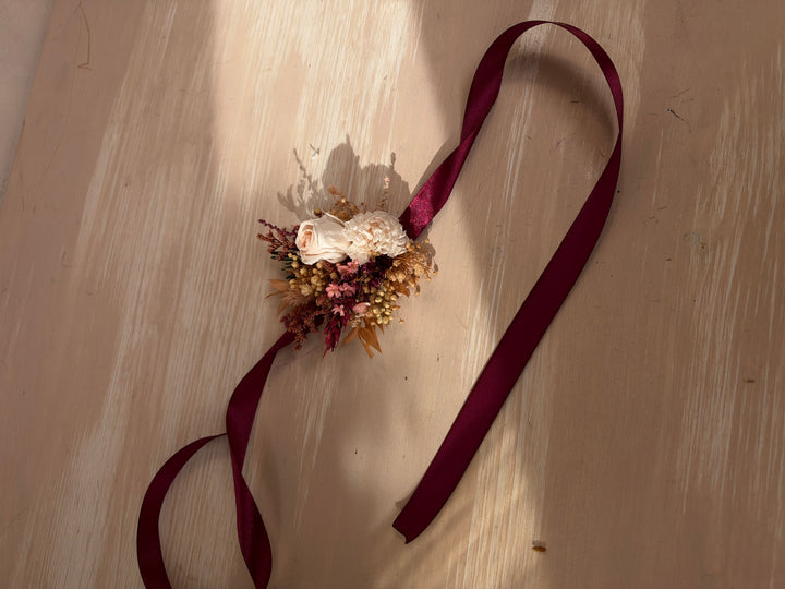 A dried floral bracelet with a long, wide burgundy ribbon, laid out on a light wood surface with soft sunlight and shadows.
