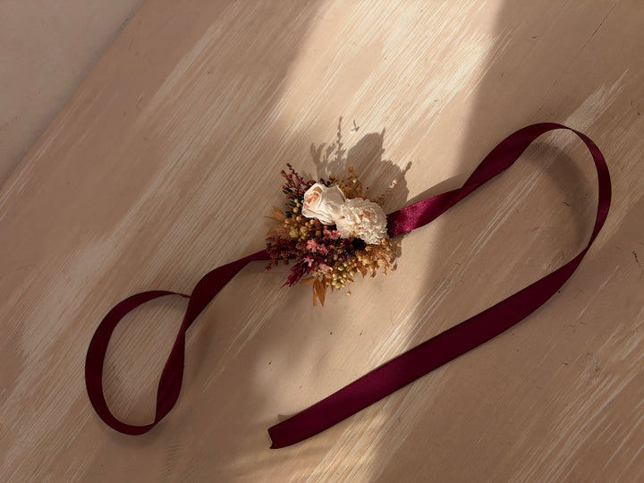 A dried floral wrist corsage with a burgundy ribbon, laid out on a light wood surface with warm sunlight and shadows.
