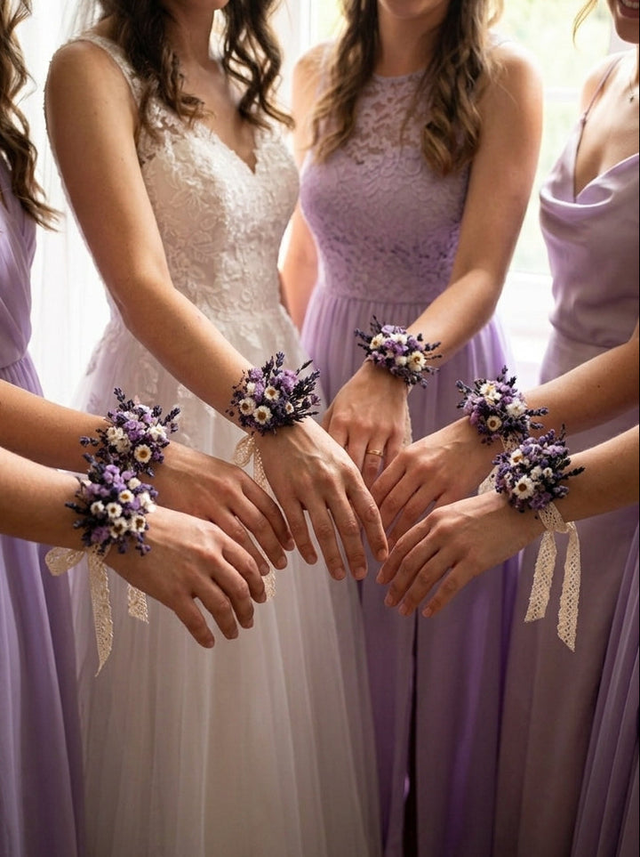 Four women standing together in formalwear, with three wearing lavender bridesmaid dresses and one in a white lace gown. Each woman has a matching wrist corsage with purple flowers and white lace ribbon, gathered in a circle at the center of their hands.