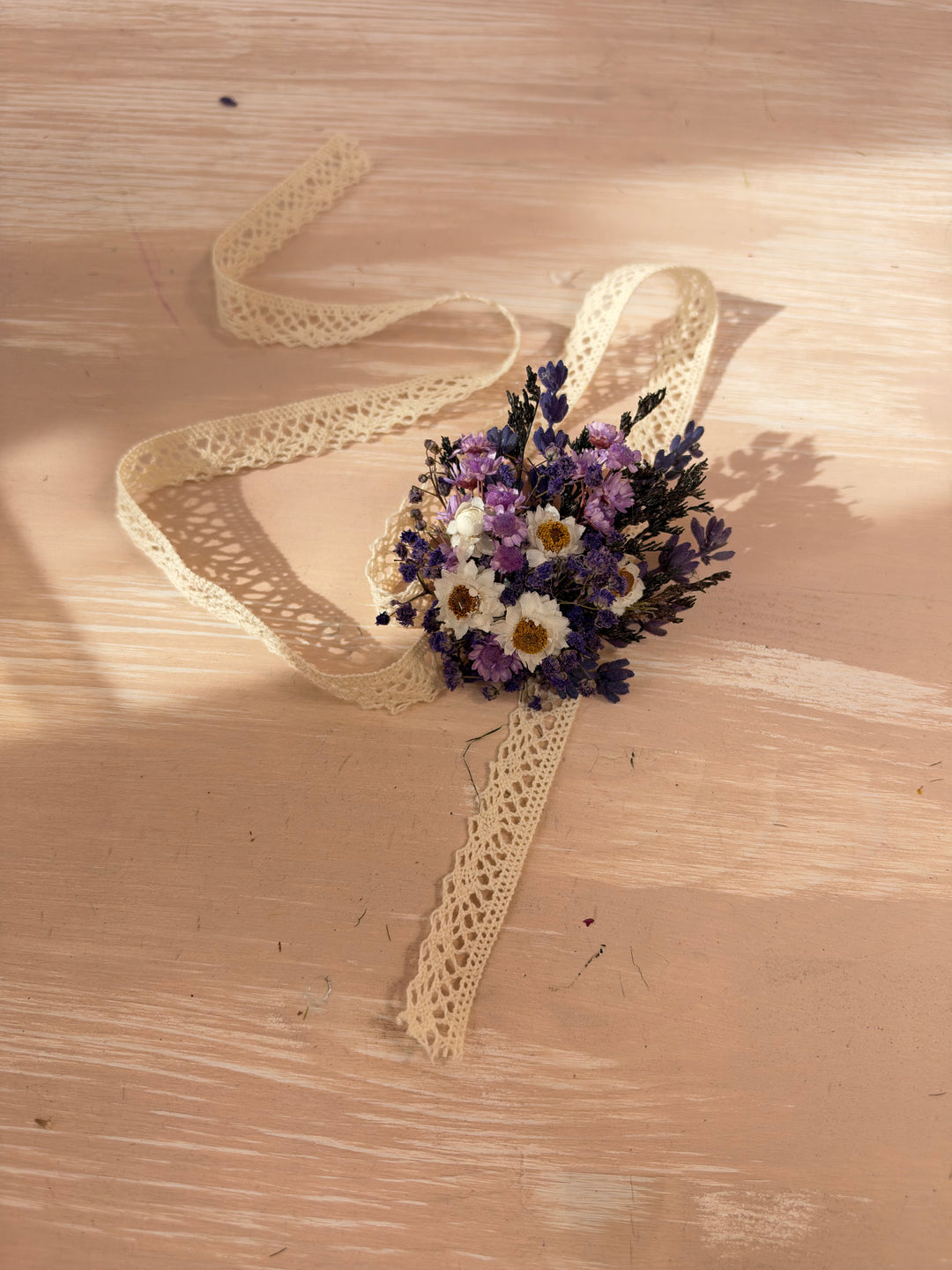 A small dried flower bracelet with purple and white blossoms tied with a cream lace ribbon, placed on a light wood surface with warm sunlight and shadows.