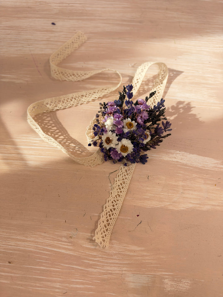 A small dried flower bracelet with purple and white blossoms tied with a cream lace ribbon, placed on a light wood surface with warm sunlight and shadows.