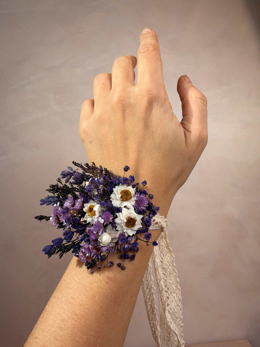 A wrist corsage featuring dried purple and white flowers with a cream lace ribbon, worn on a person's wrist against a neutral beige background.