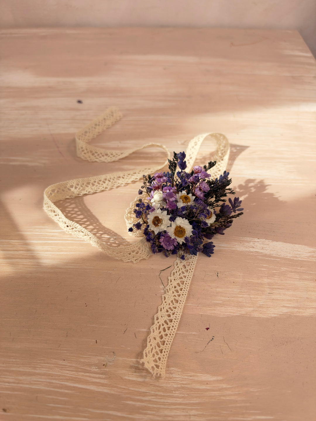 A small dried flower bracelet with purple, white, and yellow blooms tied with a long cream lace ribbon, arranged on a light wood surface with warm sunlight and shadows.