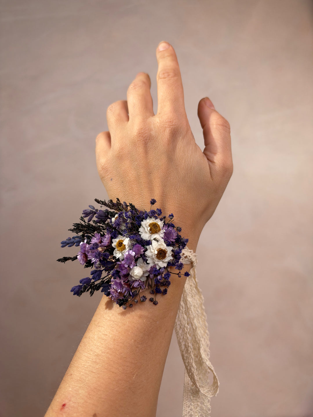 A wrist corsage featuring dried purple and white flowers with dark green leaves, tied with a cream lace ribbon, shown worn on a person's wrist against a neutral beige background.