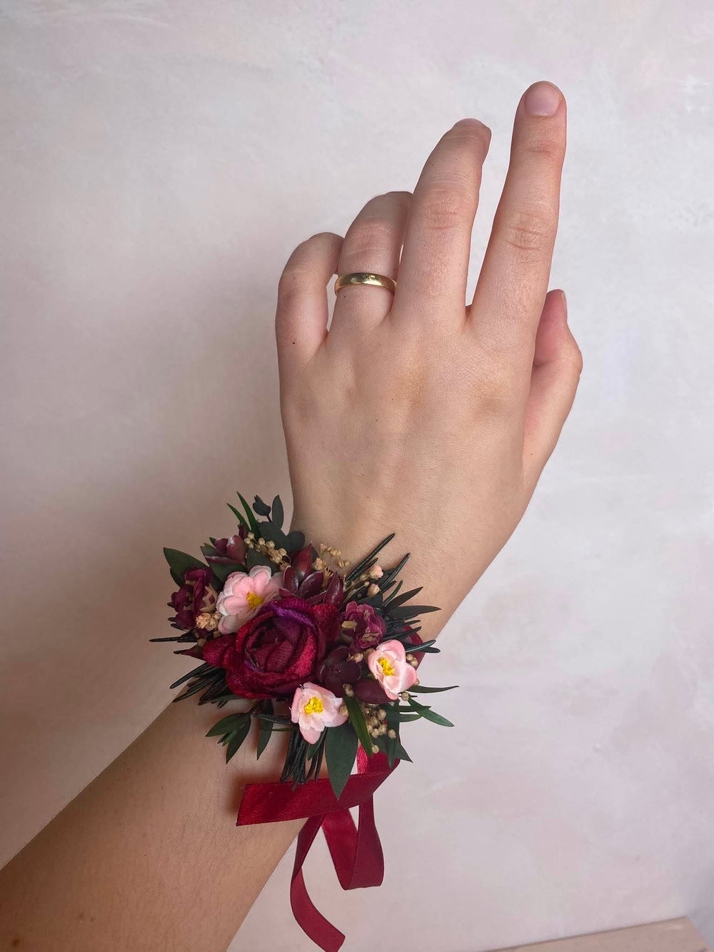 A wrist corsage featuring deep burgundy roses, light pink filler flowers, green foliage, and a burgundy ribbon, shown worn on a person's wrist against a light beige background.