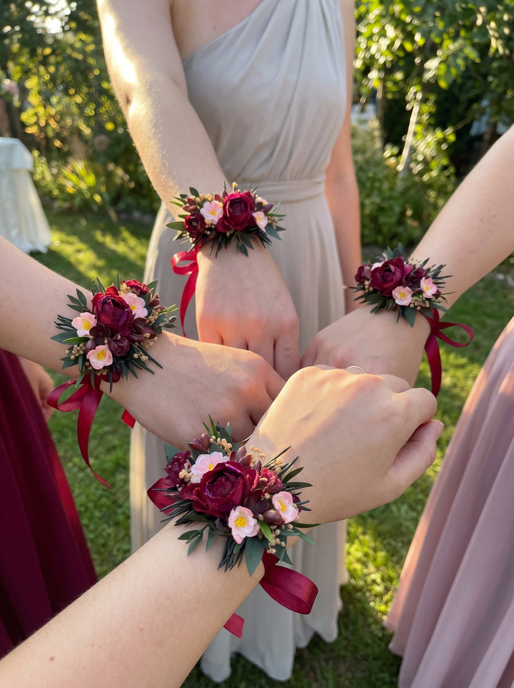 Four bridesmaids' hands gathered in the center, each wearing a matching floral wrist corsage with burgundy roses, green foliage, and burgundy ribbons. The background shows a sunny outdoor garden with green grass and trees, and the bridesmaids are wearing light-colored dresses.