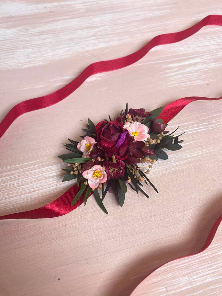 A floral wrist corsage featuring burgundy and pink flowers with green foliage, tied with a long red satin ribbon on a light wood background.