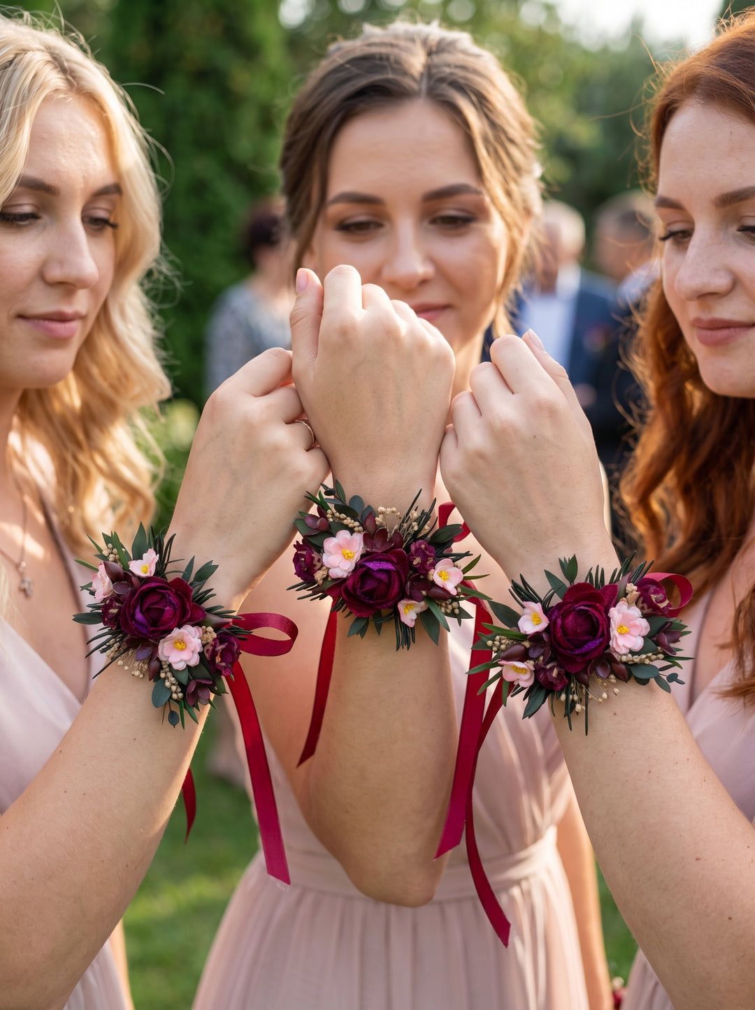 Three bridesmaids outdoors wearing matching floral wrist corsages with burgundy roses, green foliage, and red ribbons, paired with blush pink dresses.